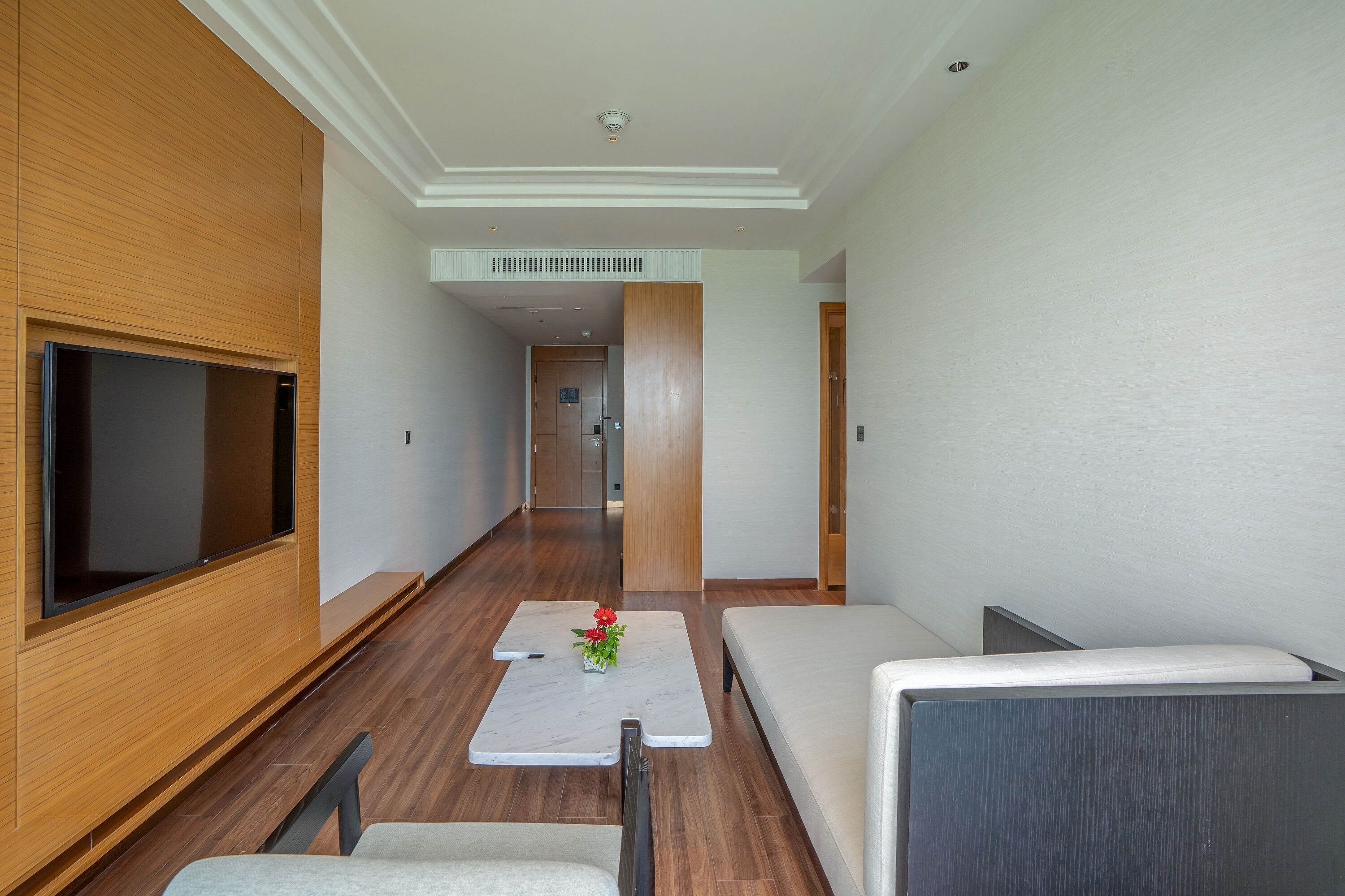 A modern hotel room with a flat-screen TV, a sofa, a small table with a vase of flowers, and a wooden panel wall.