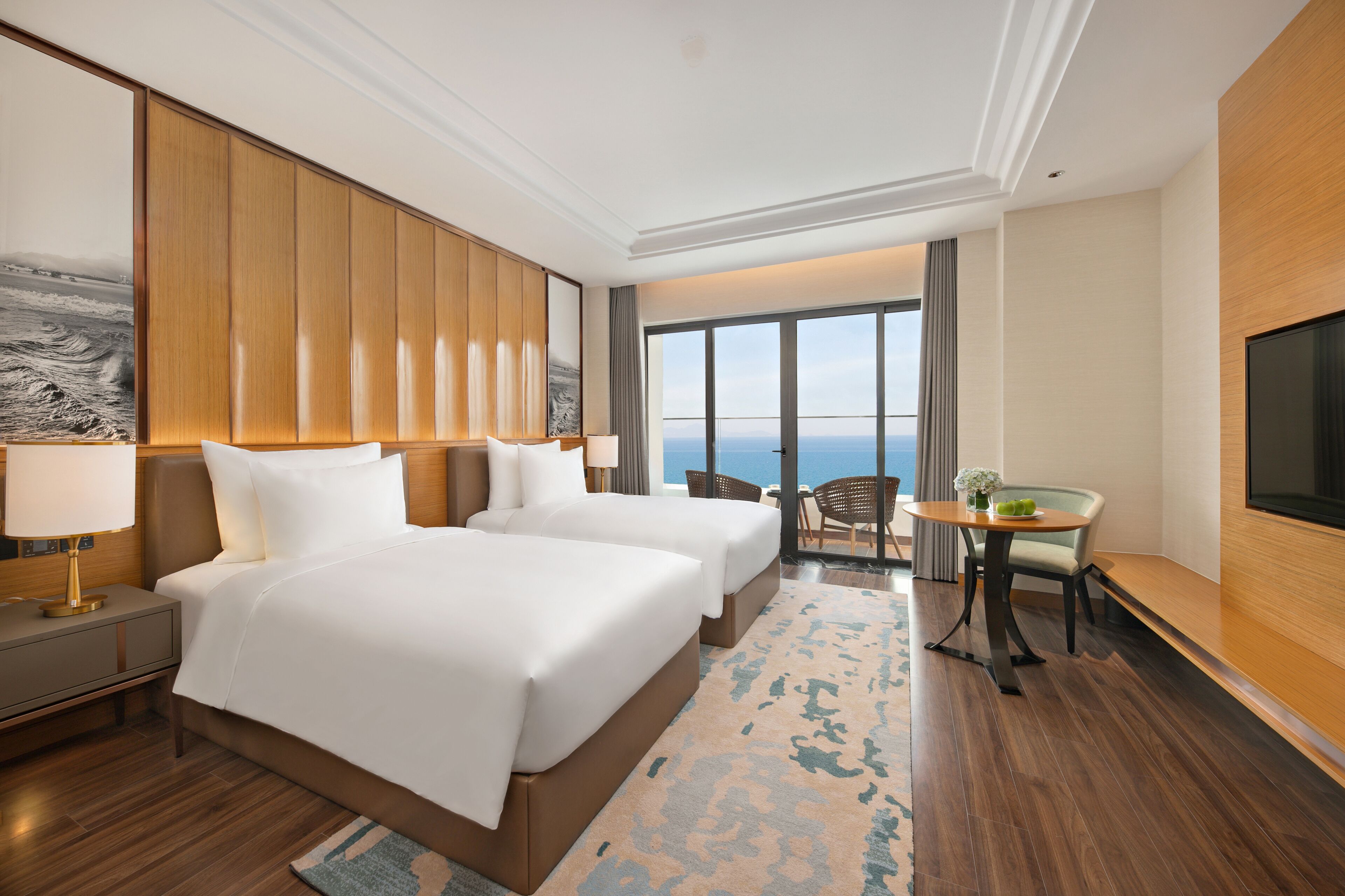 A hotel room with two beds, a desk, a chair, a TV, and a large window with a view of the ocean.