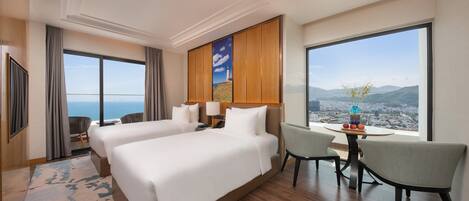 Habitación de hotel con dos camas, un comedor y un amplio ventanal con vistas a la ciudad y a la montaña.