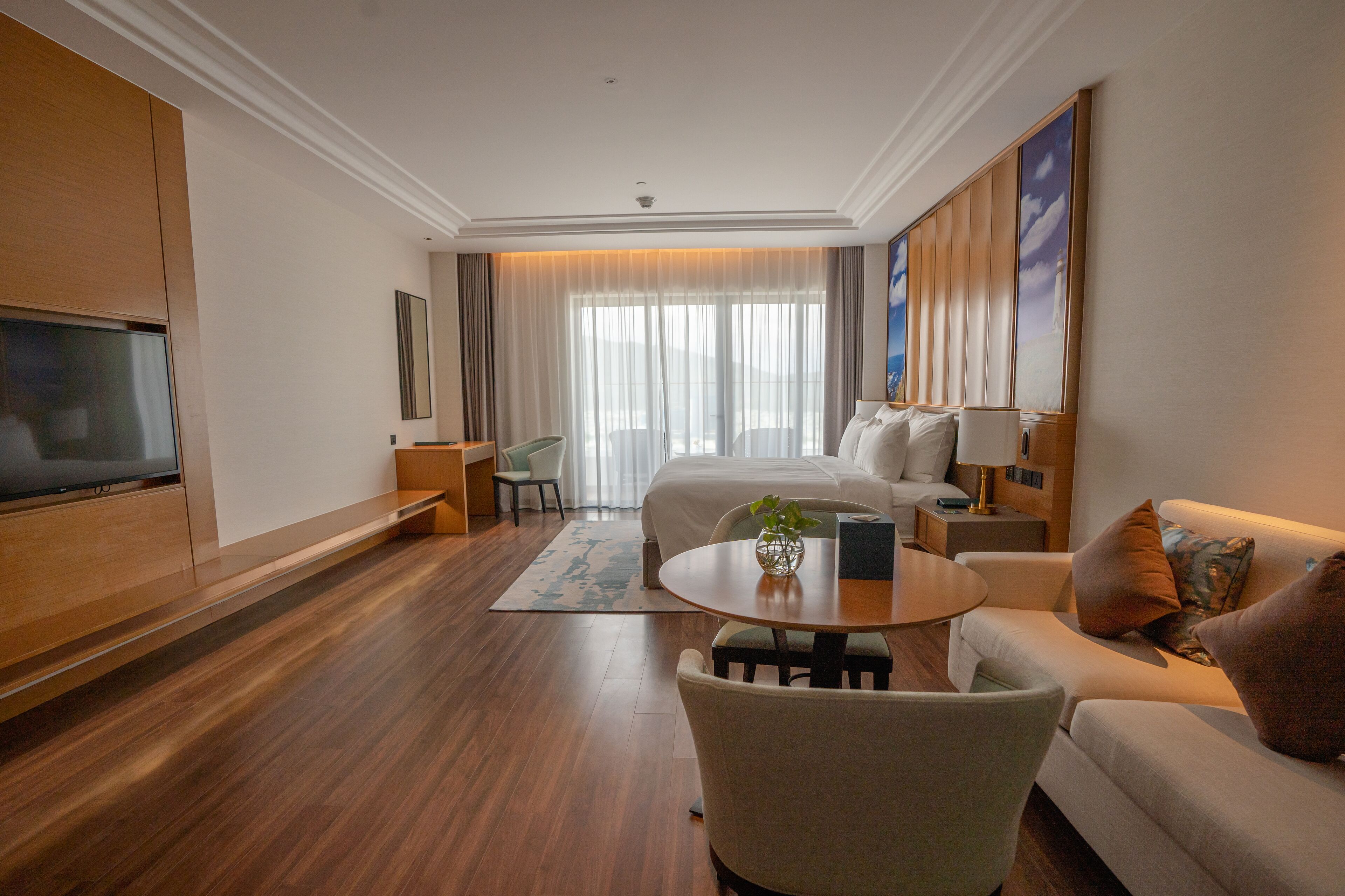 A modern hotel room with a flat-screen TV, a sofa, a bed, a dining table, and a large window with curtains.