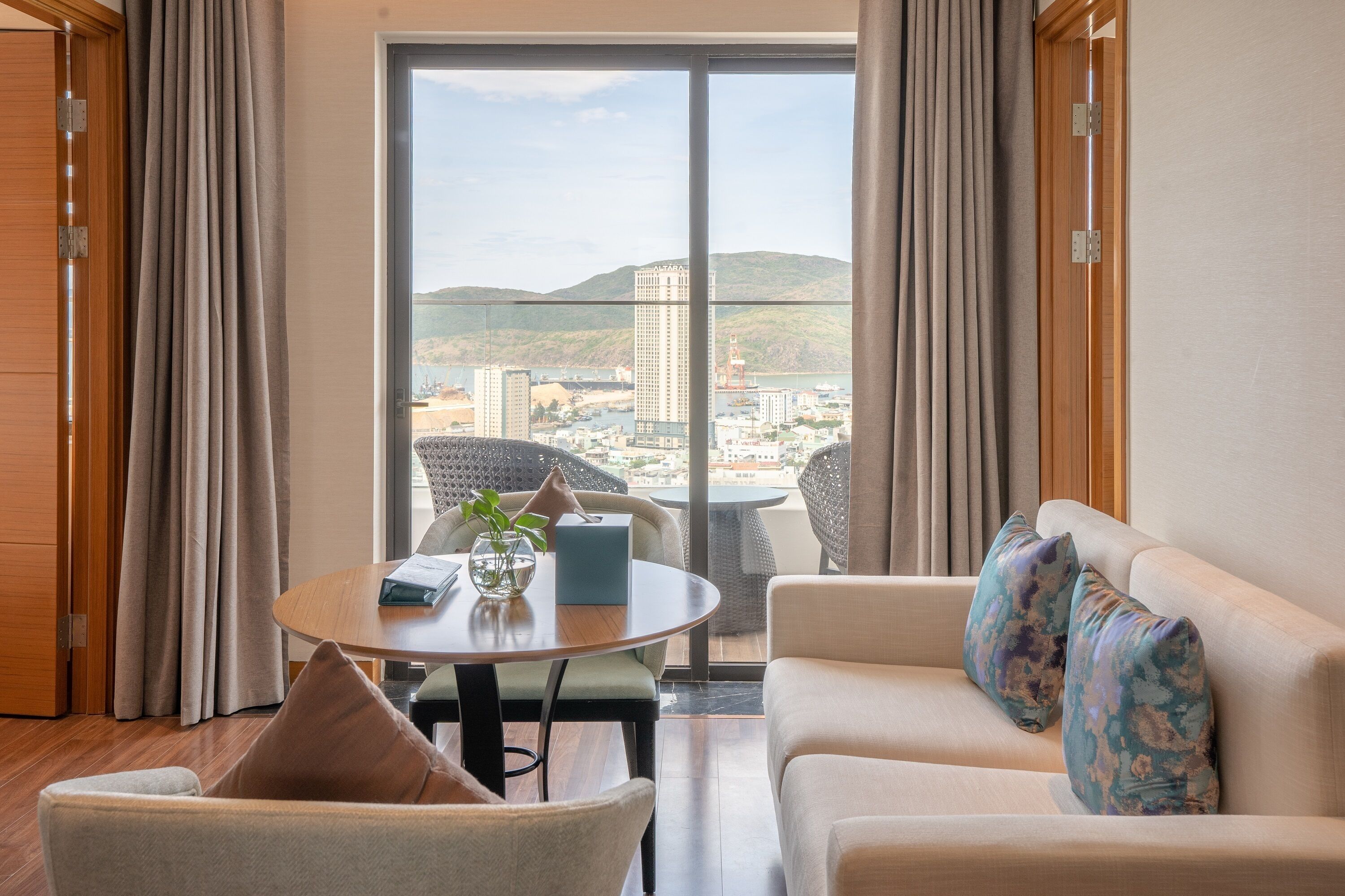 A modern hotel room with a large window, a round dining table, a sofa, and a chair.