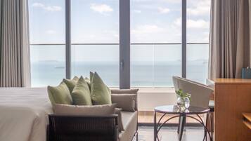 A hotel room with a large window offering a view of the sea. There is a bed with pillows, a small table with a vase, and a chair.
