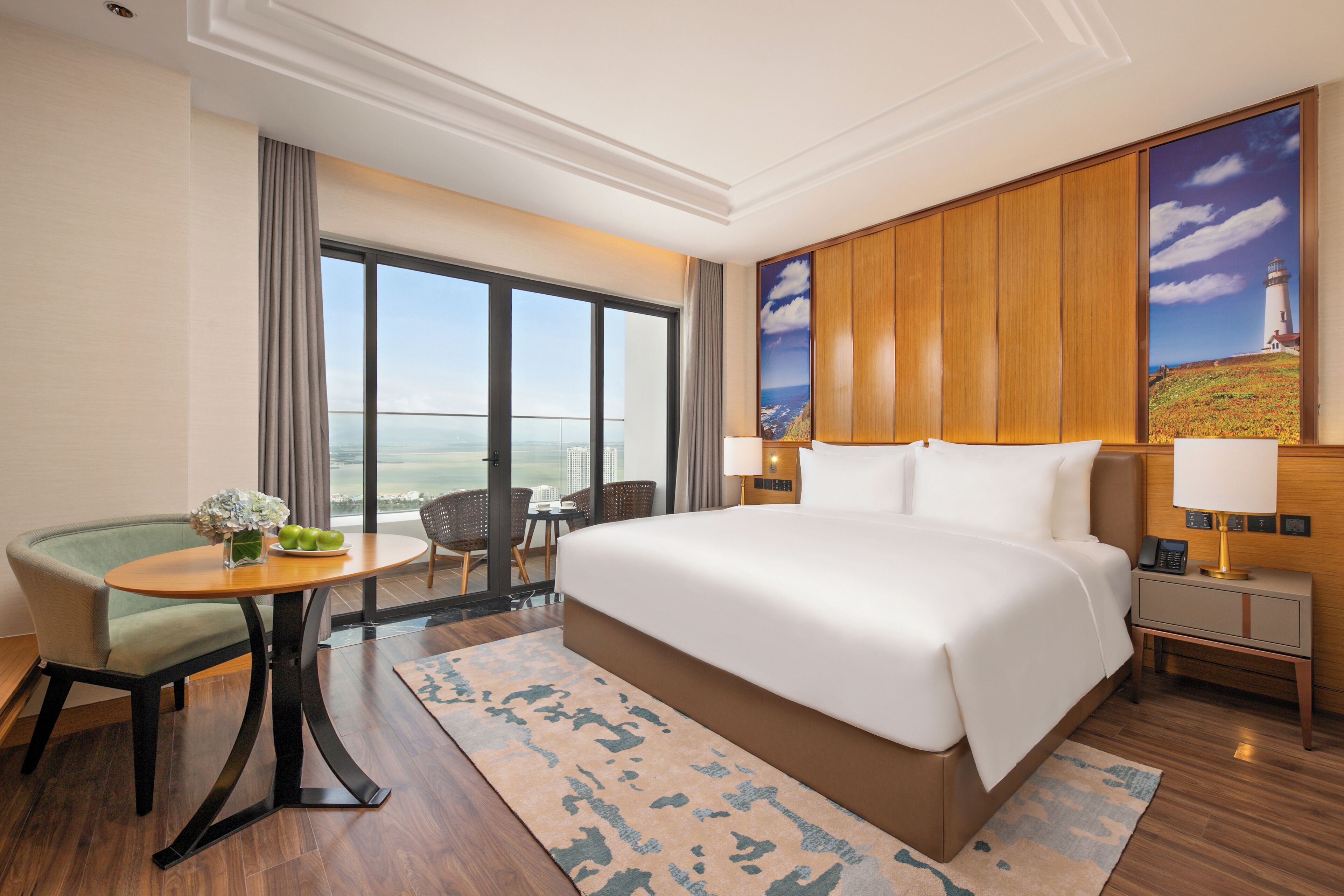 A hotel room with a large bed, a dining table, a chair, a small table with a vase, and a painting of a lighthouse.