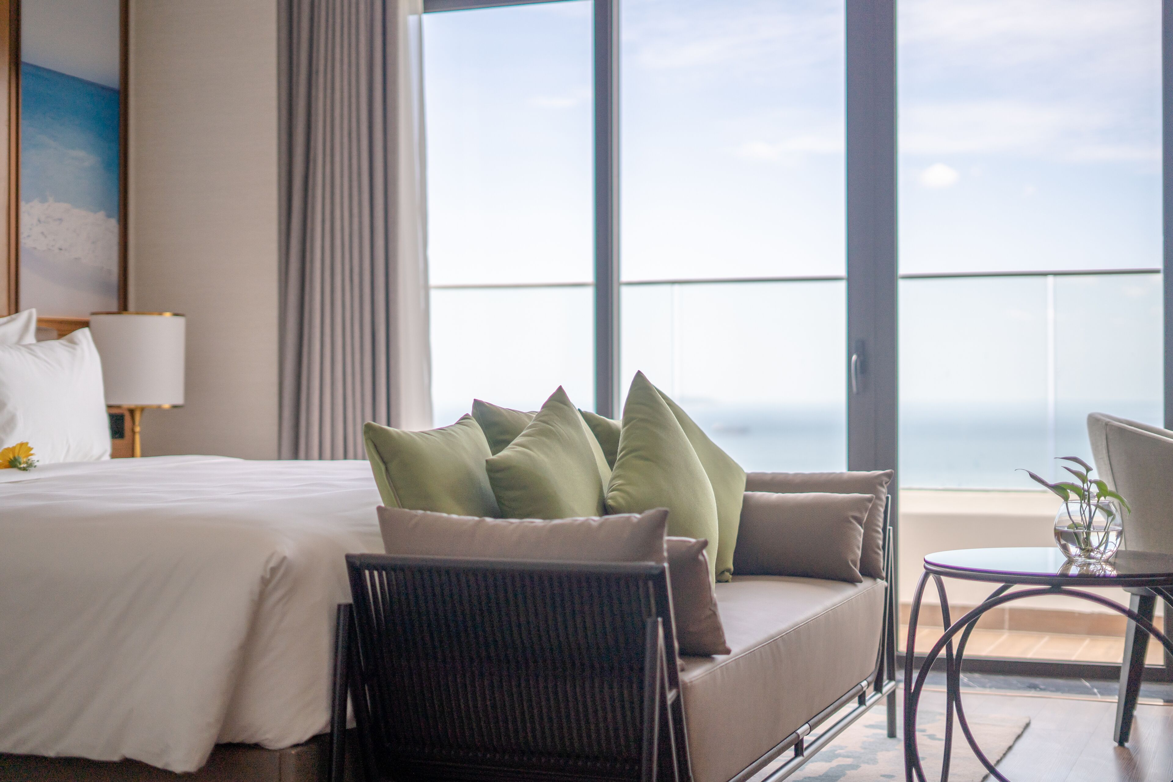 Une chambre d’hôtel comprenant un lit, un canapé avec des coussins, une chaise, une petite table avec un vase et une grande fenêtre donnant sur le ciel et les nuages.