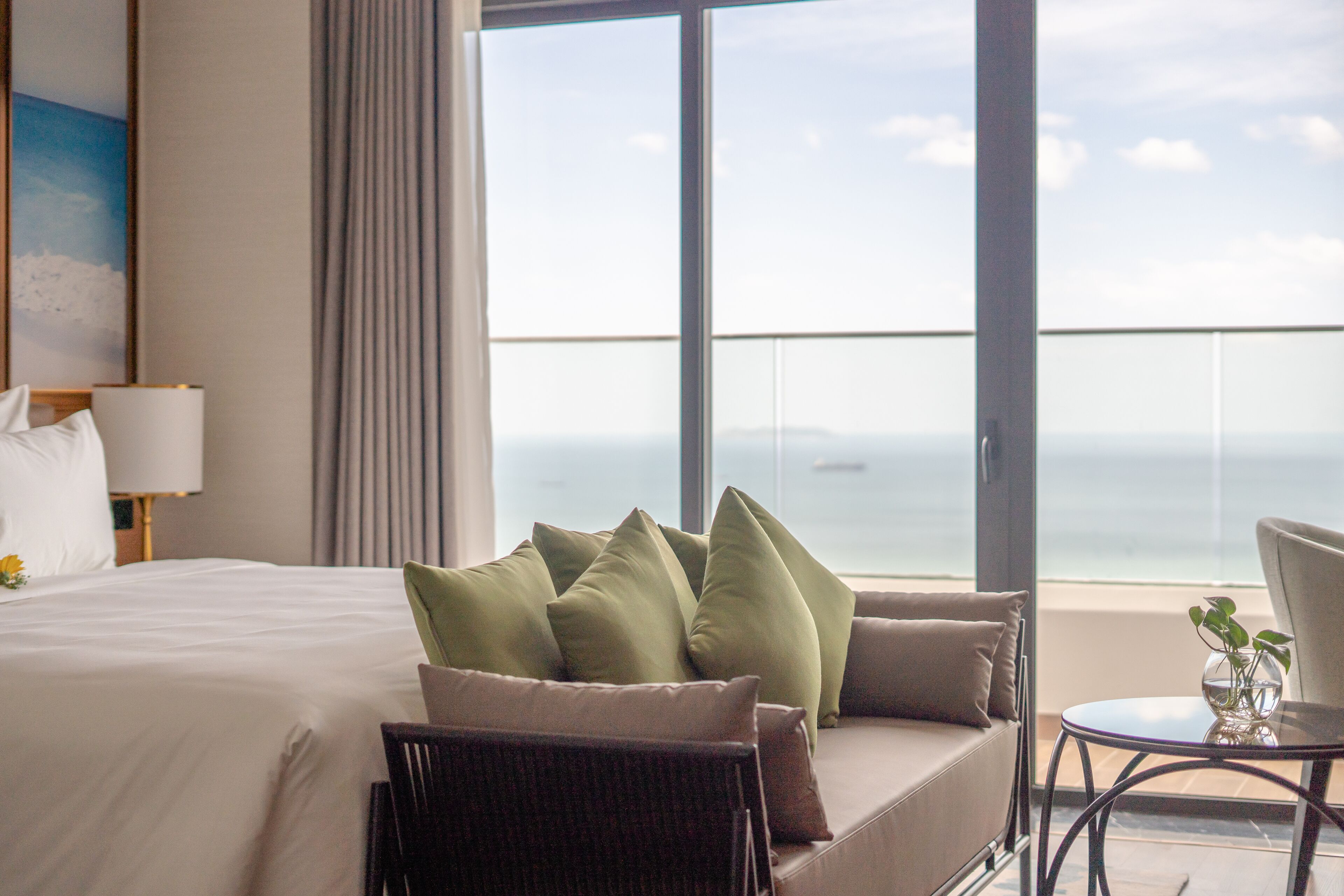 Une chambre d’hôtel avec un grand lit, un canapé, un fauteuil et une petite table. De grandes fenêtres offrent une vue sur la mer.