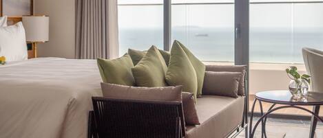 Une chambre d’hôtel avec un grand lit, un canapé, un fauteuil et une petite table. De grandes fenêtres offrent une vue sur la mer.