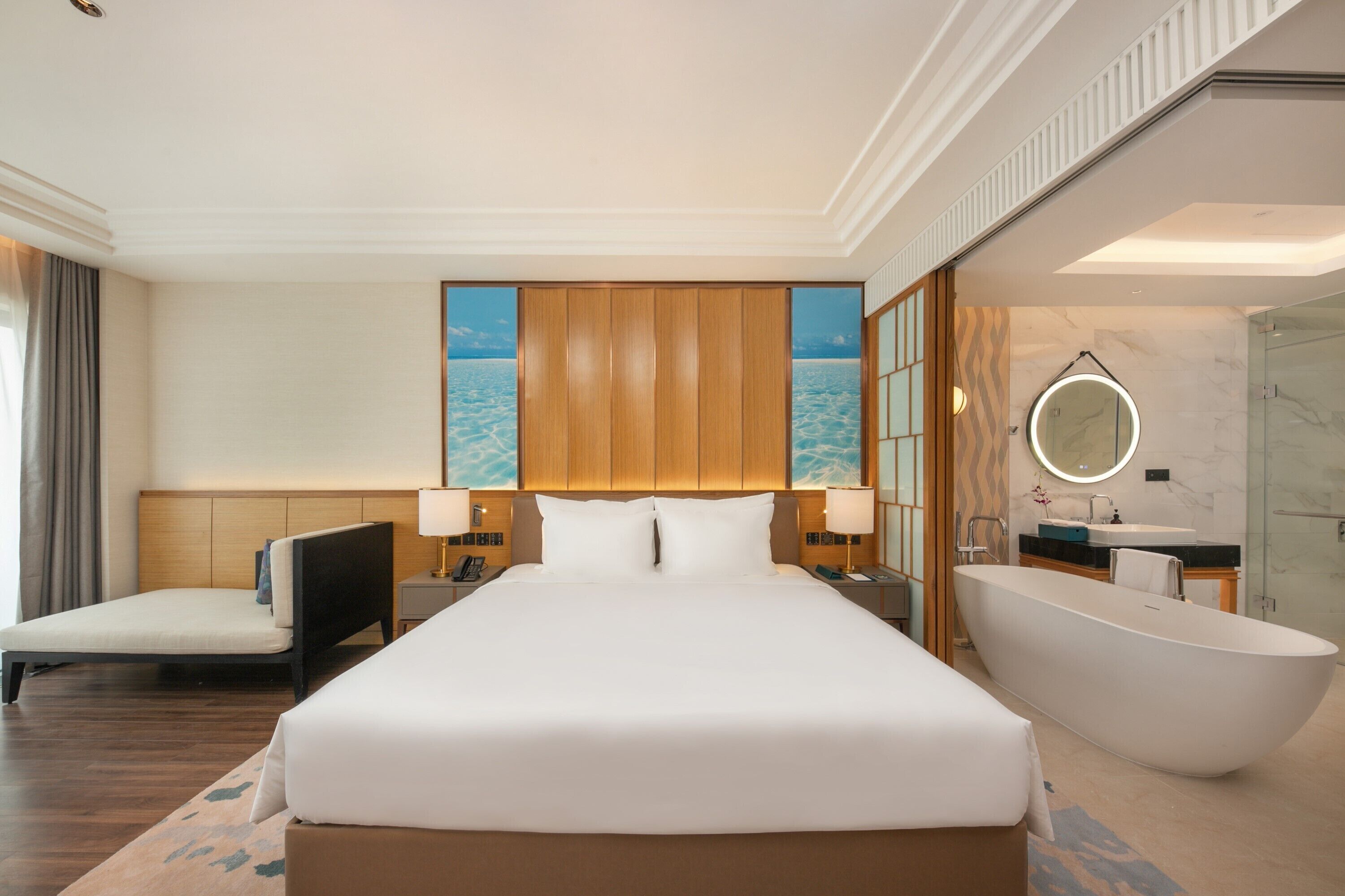 A hotel room with a large bed, a sitting area, a bathtub, and a bathroom with a marble wall and a round mirror.
