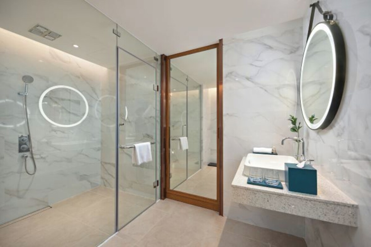 A modern bathroom with a glass shower enclosure, a wooden door, a marble wall, a round mirror, and a countertop with a sink and toiletries.