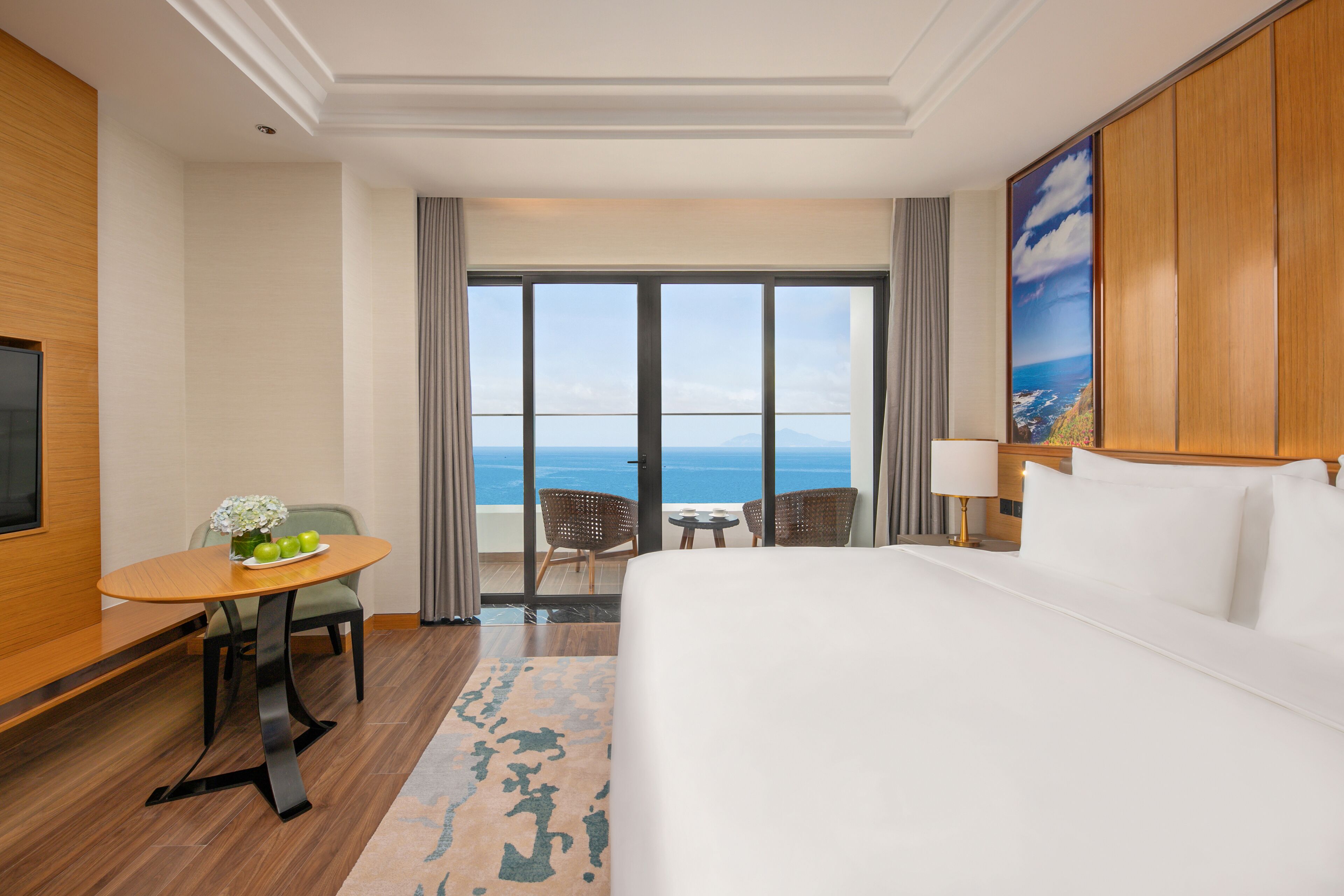 A hotel room with a large bed, a dining table with chairs, a small table with a lamp, and a view of the ocean through large windows.
