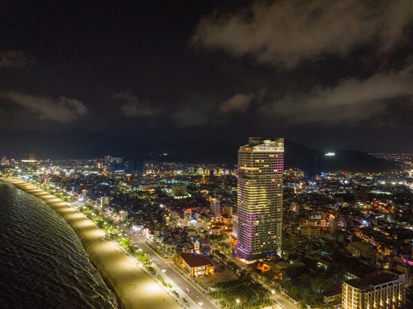 A nighttime cityscape with a prominent illuminated skyscraper, a beach, and a mountainous backdrop. - Grand Hyams Hotel - Quy Nhon Beach (Quy Nhon)
