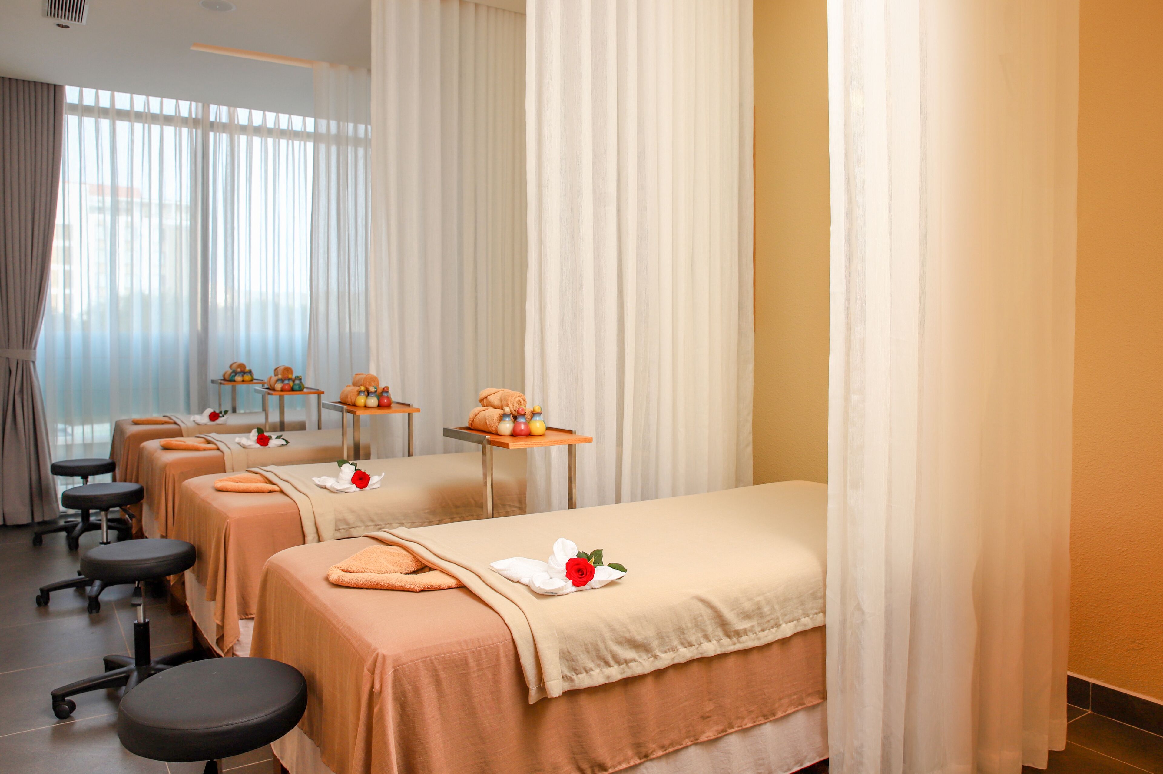Une salle de spa équipée de trois tables de massage, chacune agrémentée d’une serviette et d’une rose. On y trouve également des chaises et une petite table sur laquelle sont disposés des fruits et un vase.