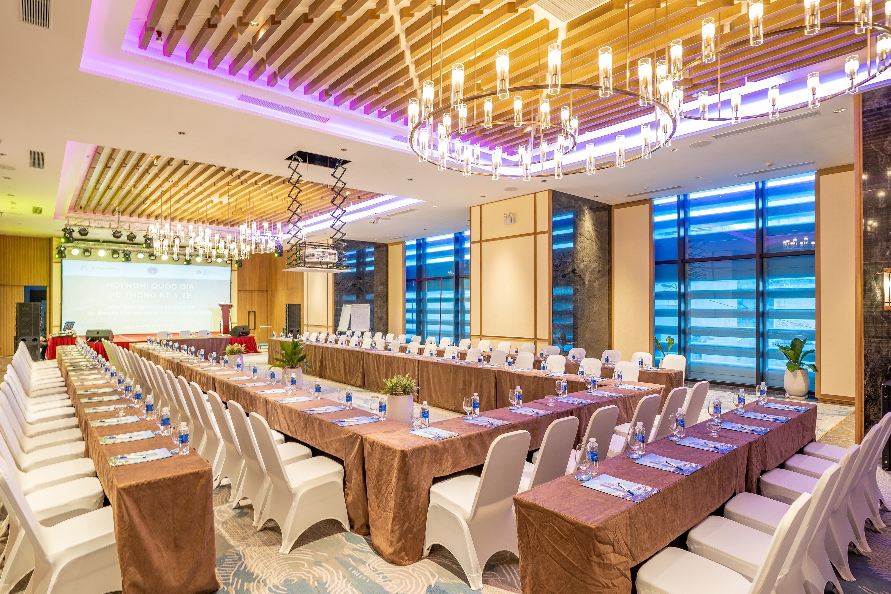 A conference room with a long table, white chairs, and a large screen at the front.
