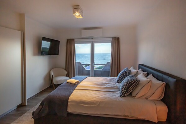 A hotel room with a bed, a chair, a TV, and a view of the sea.
