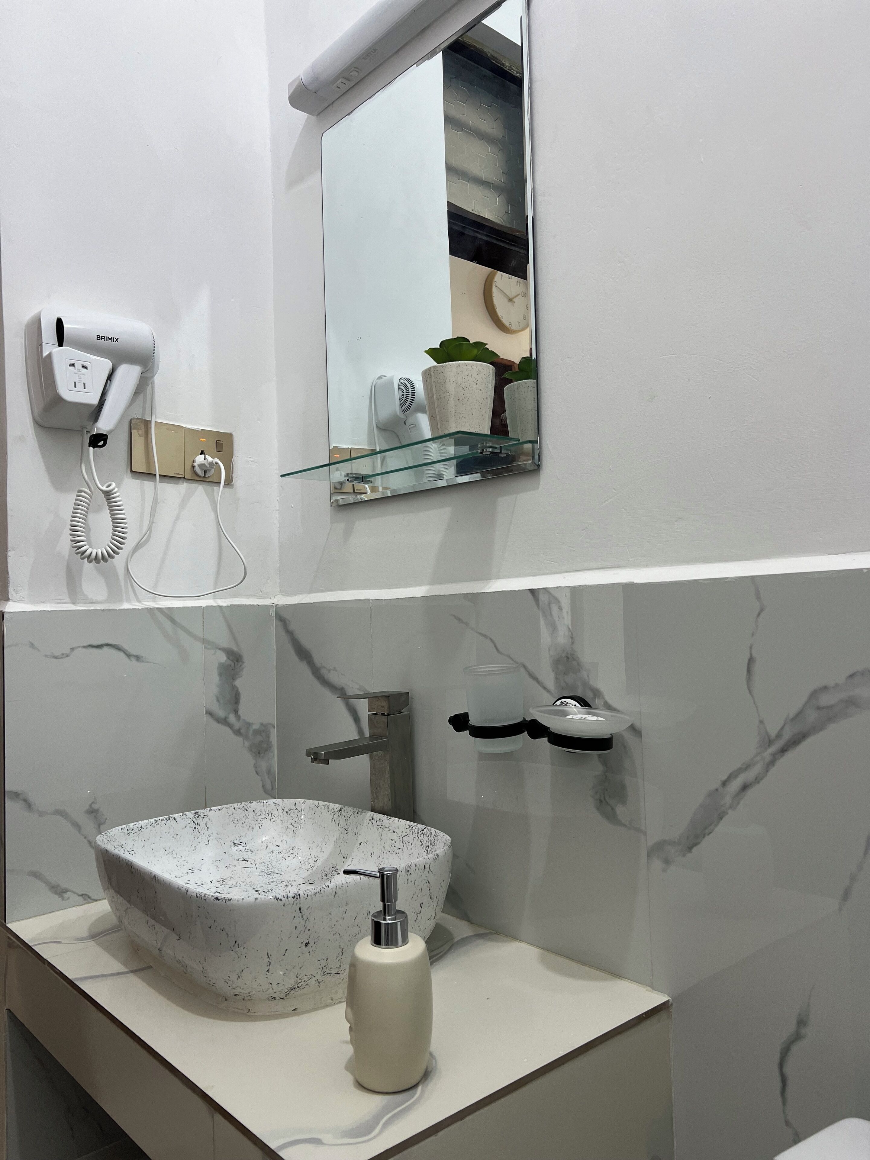 A bathroom with a marble sink, a handheld hair dryer, a glass shelf with toiletries, and a wall-mounted mirror.