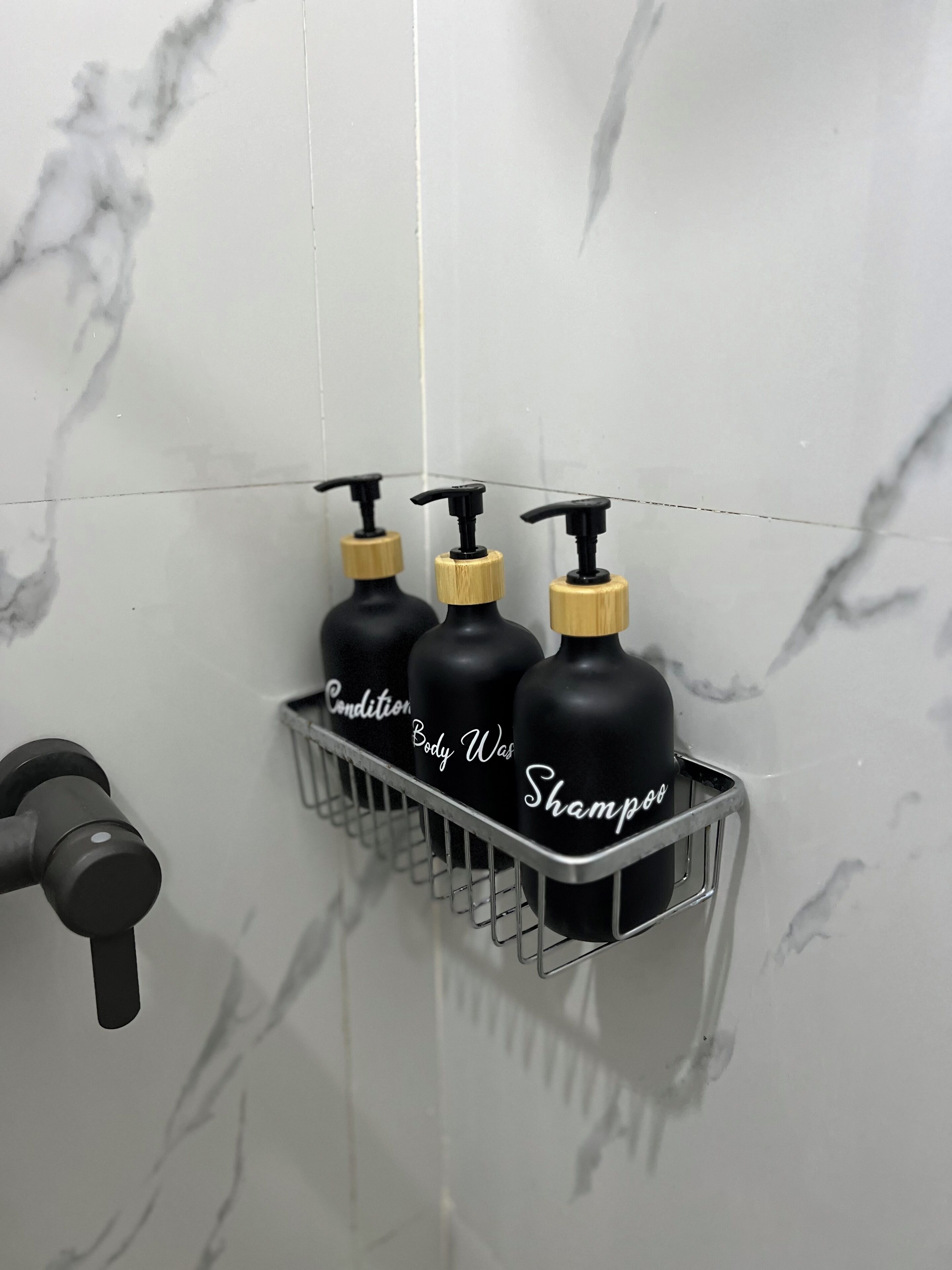 A bathroom with three black shampoo bottles in a metal rack, a black faucet, and a marble wall.