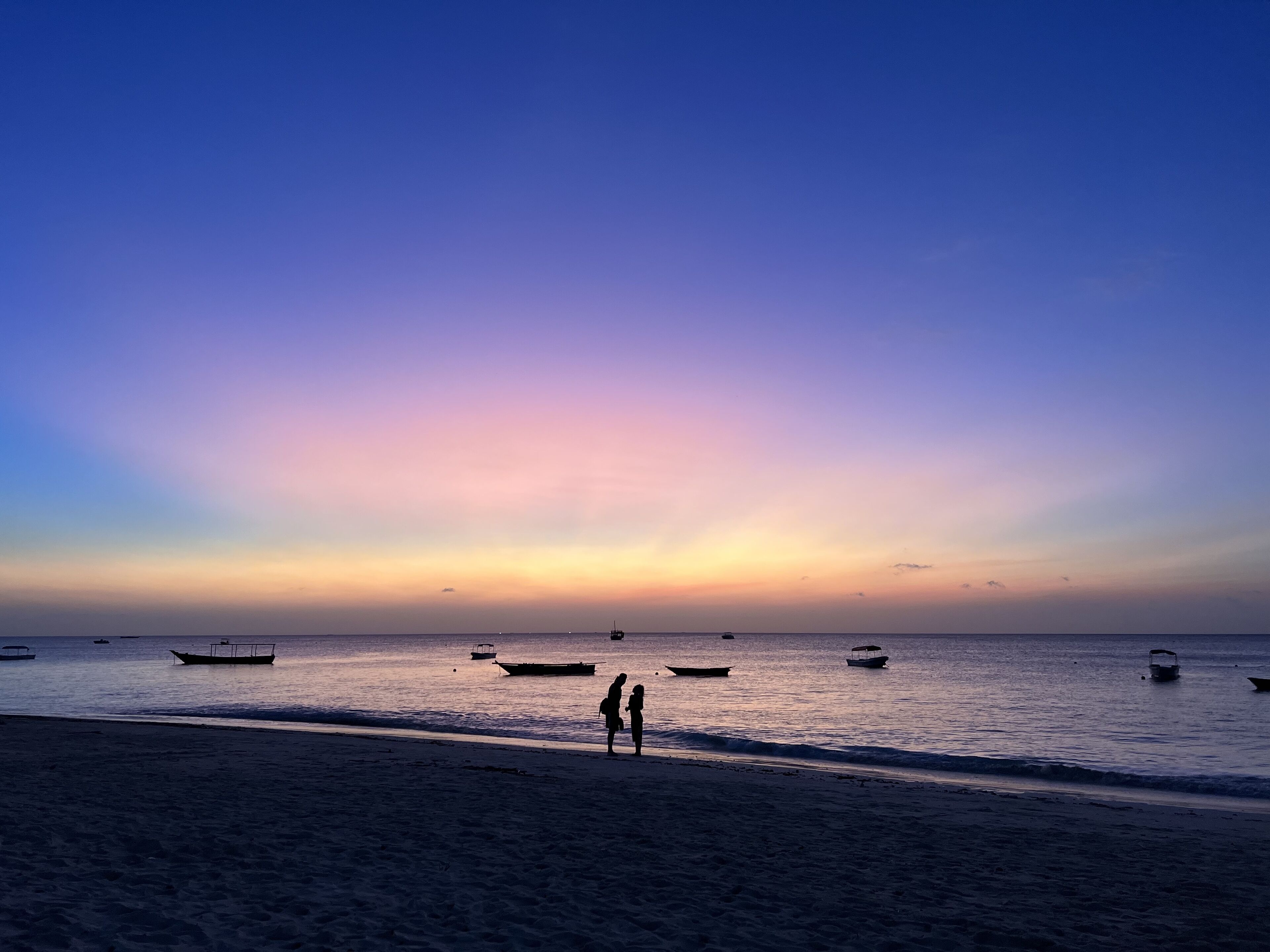 A beach with boats in the water and two people standing on the sand.