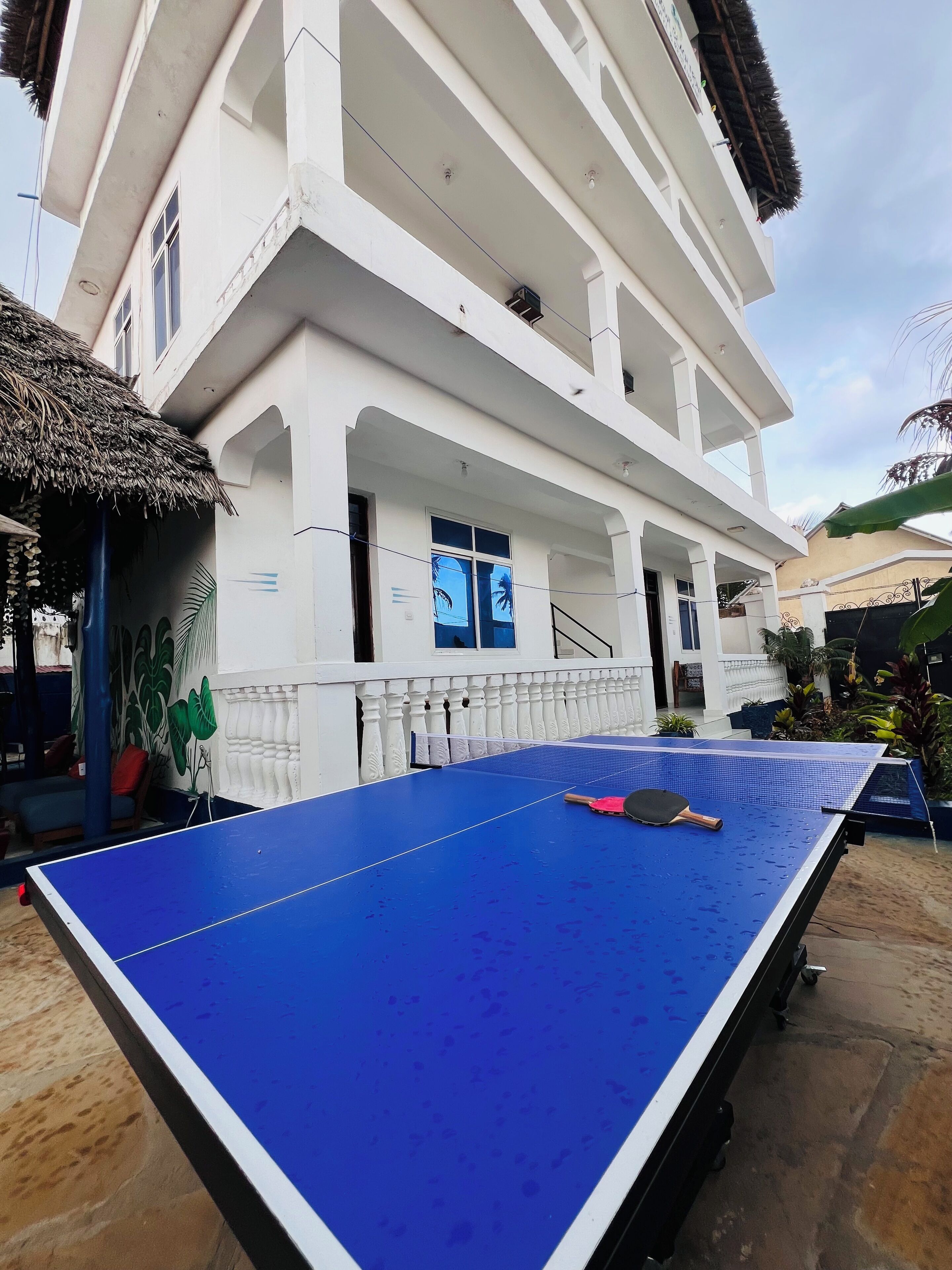 Une table de ping-pong bleue, avec des raquettes, est installée à l’extérieur, devant un bâtiment blanc doté de balcons et d’une structure à toit de chaume située à proximité.