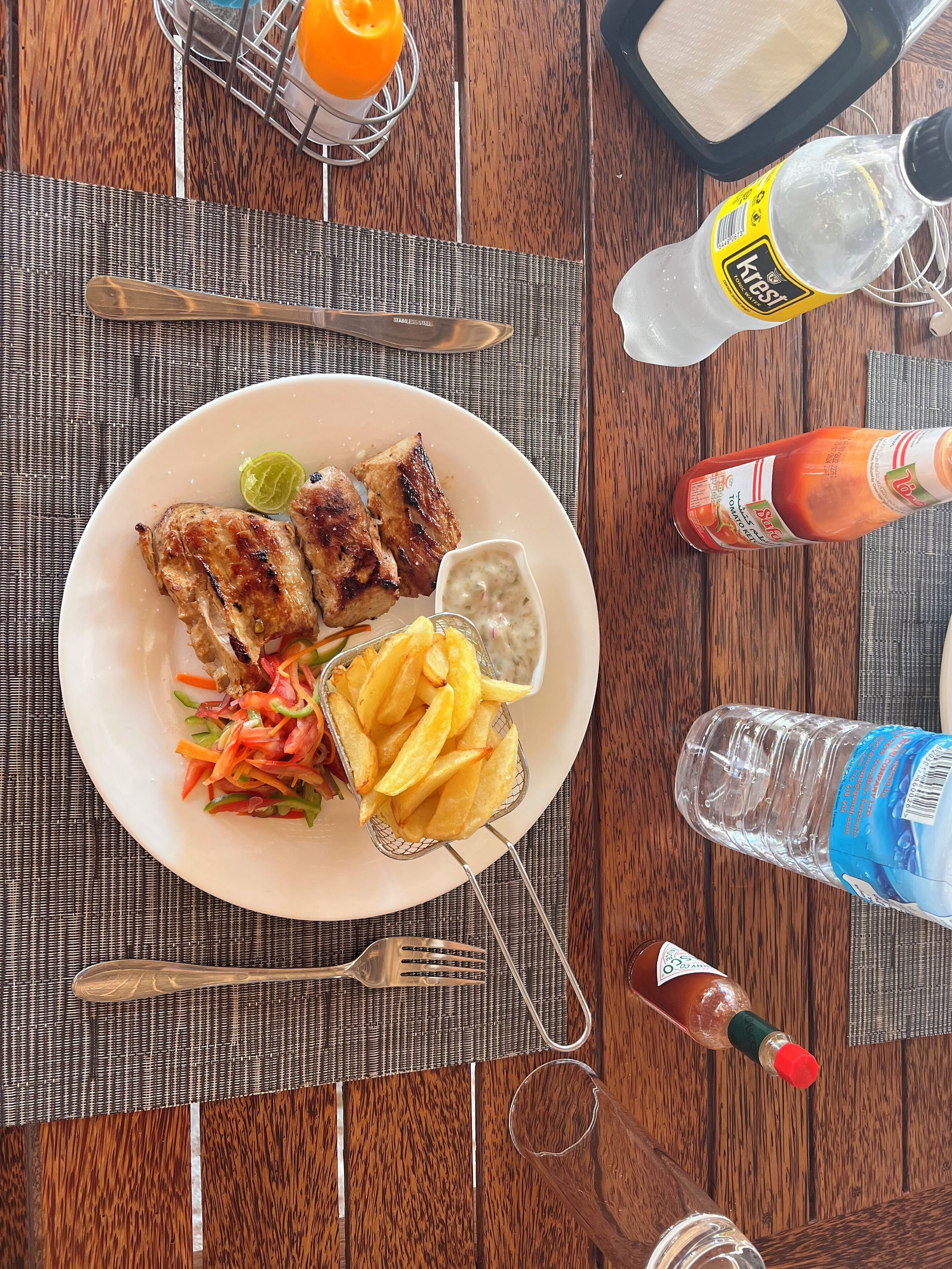 A dining table with a plate of food, including grilled chicken, fries, and a salad, along with condiments and a bottle of water.