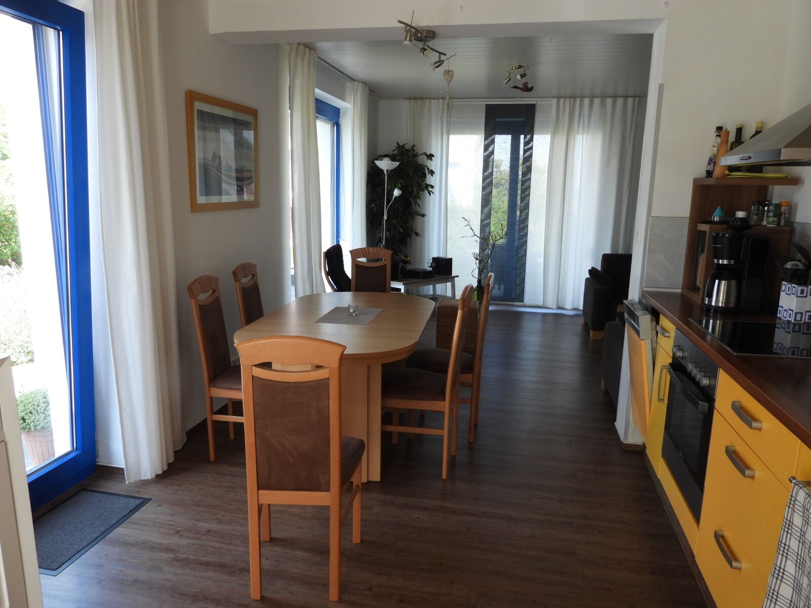 A dining area with a round table and six chairs, a kitchen with yellow cabinets, and a large window with curtains.