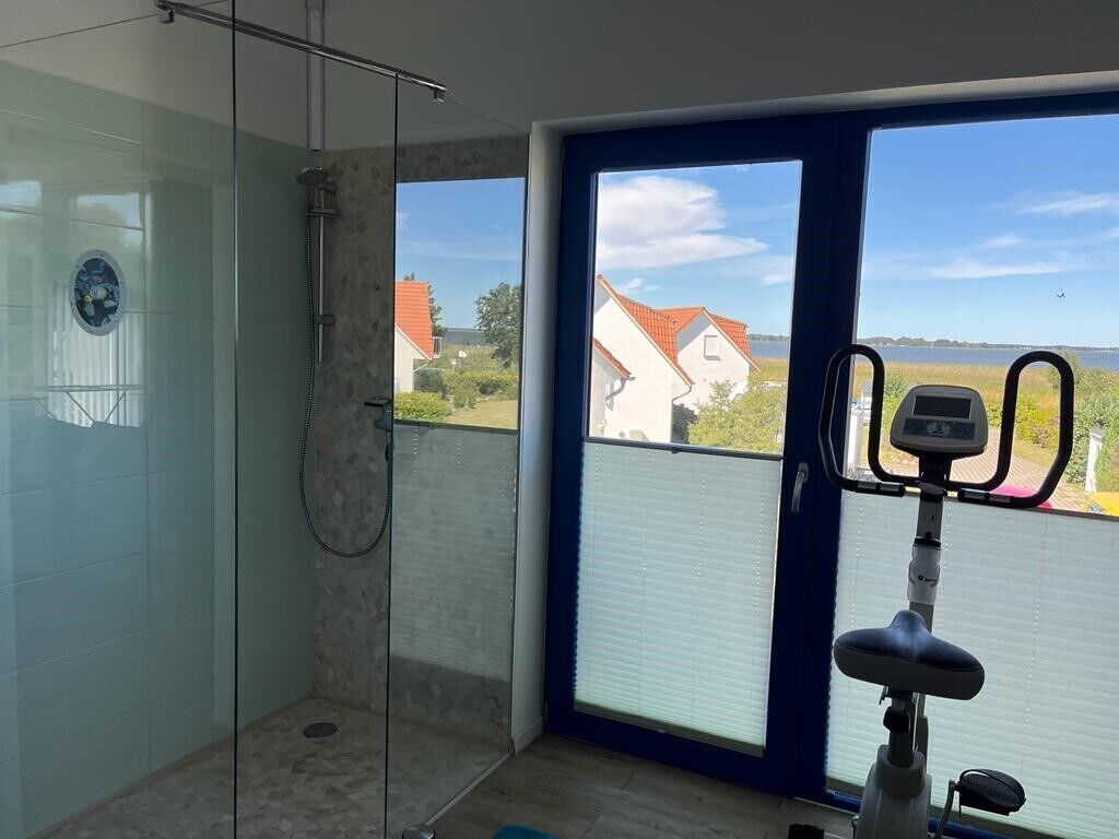 A bathroom with a glass shower enclosure, a window with a view of houses and a body of water, and a stationary exercise bike.