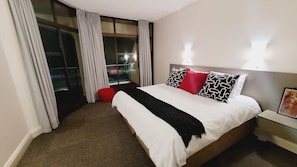 A hotel room with a large bed, a red ottoman, and a view of the outdoors through a sliding glass door. - Effortless Homes (East London)