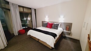 A hotel room with a large bed, a red ottoman, a glass door leading to a balcony, and a nightstand with a framed picture. - Effortless Homes (East London)