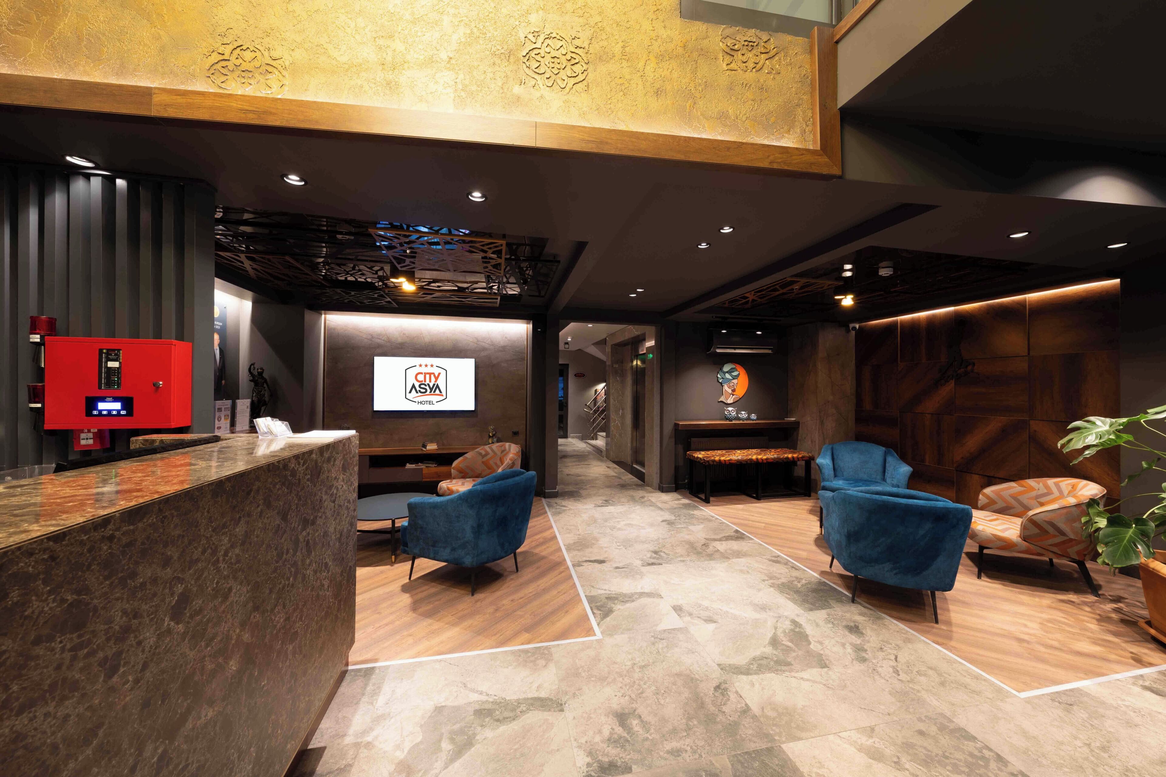 A hotel reception area with a reception desk, blue armchairs, a red fire alarm, and a wall-mounted ASYA HOTEL sign.