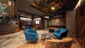 A modern interior with a reception desk, blue armchairs, a patterned ottoman, and a wall-mounted screen.