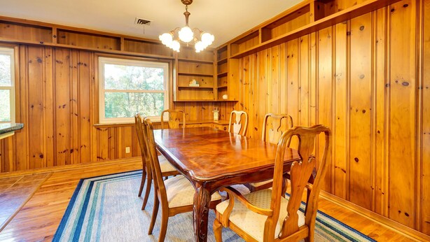 Une salle à manger aux murs lambrissés, avec une grande table et des chaises, un lustre et des étagères intégrées.