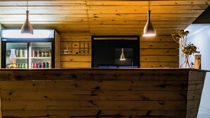 A wooden bar area with hanging pendant lights, a refrigerator stocked with beverages, and a window with blinds. - Stofpad Namibia (Solitaire)