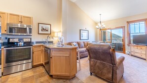 A modern kitchen with a central island, wooden cabinets, stainless steel appliances, and a dining area with a brown leather couch and a flat-screen TV. - Top Floor w/ Mountain & Ski Slope Views, Ideal Location in River Run Village (Keystone)