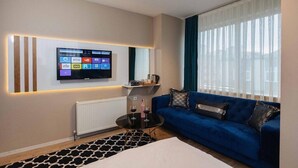 A modern hotel room with a large flat-screen TV mounted on the wall, a blue sofa, a small round coffee table, and a patterned rug. - Lina Suit Hotel (Istanbul)