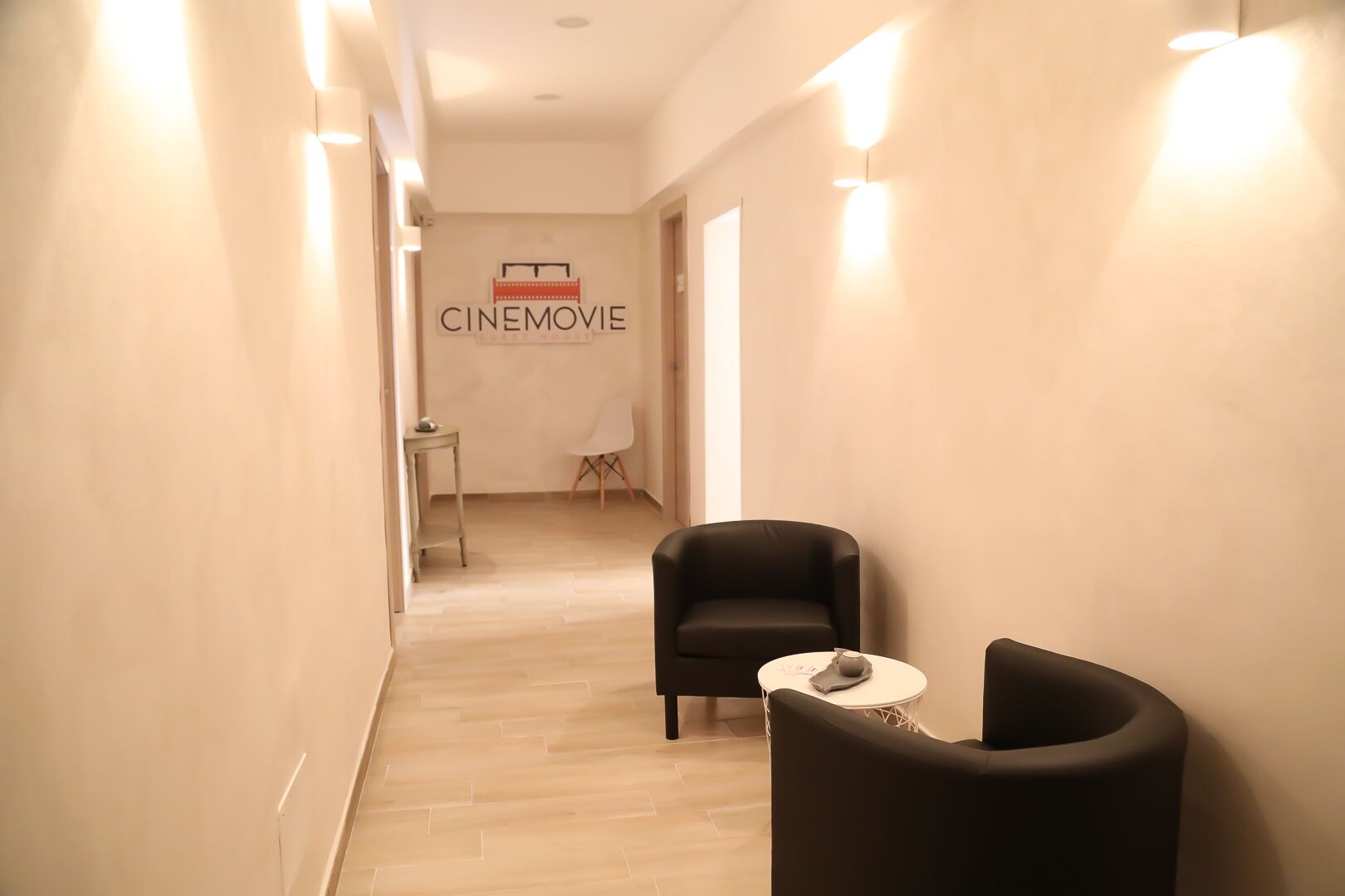 A hallway with a sign reading "CINEMOVIE" on the wall, two black chairs, a small round table, and a white chair.