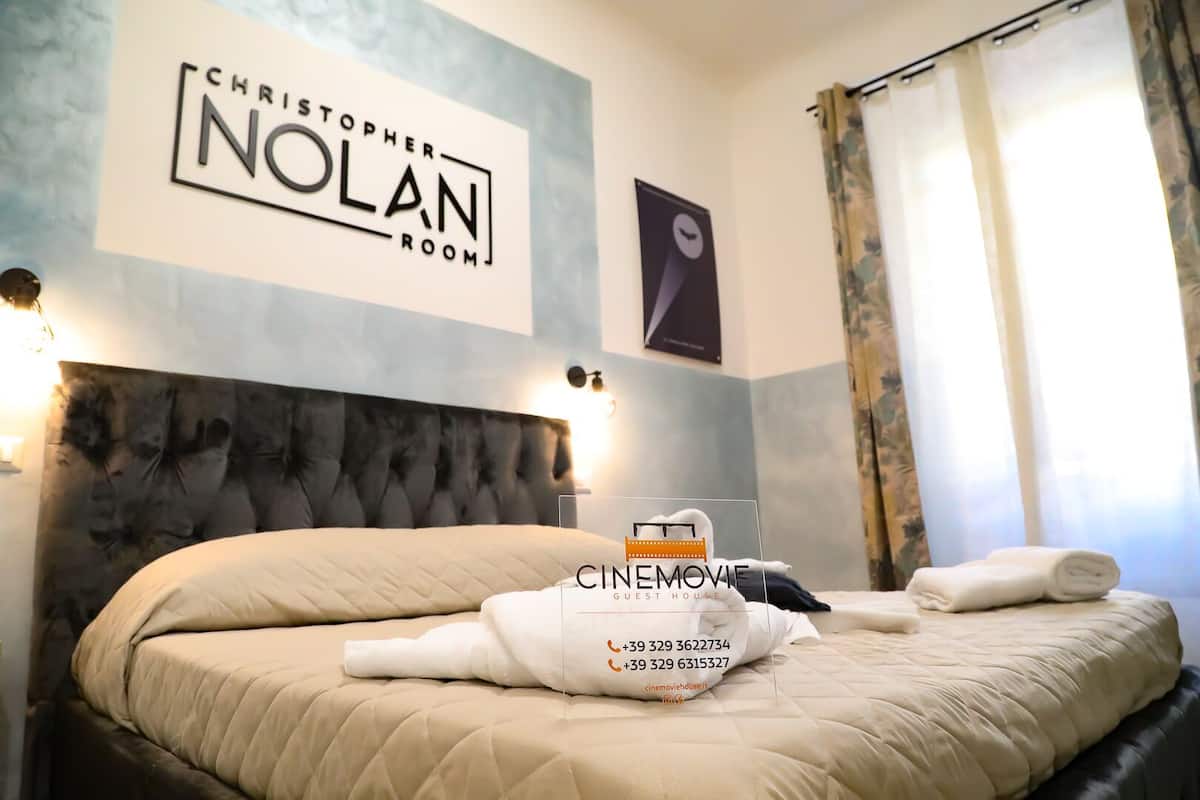 Una habitación de hotel con un cabecero capitonado, una cama con ropa de cama color beige y un cartel sobre la cama que dice 'Christopher Nolan Room'.