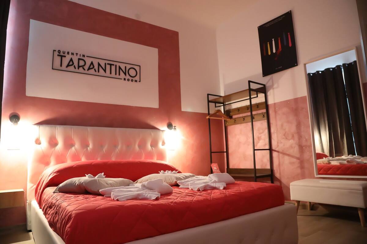 Habitación de hotel con cama roja, cabecero con luces, armario y un cartel que dice 'QUENTIN TARANTINO ROOM'.