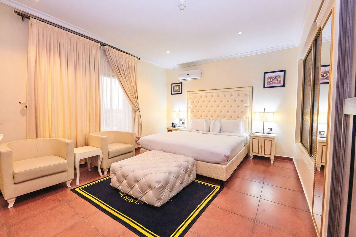 A hotel room with a large bed, two armchairs, a small side table, a bedside table with a lamp, and a large mirror.