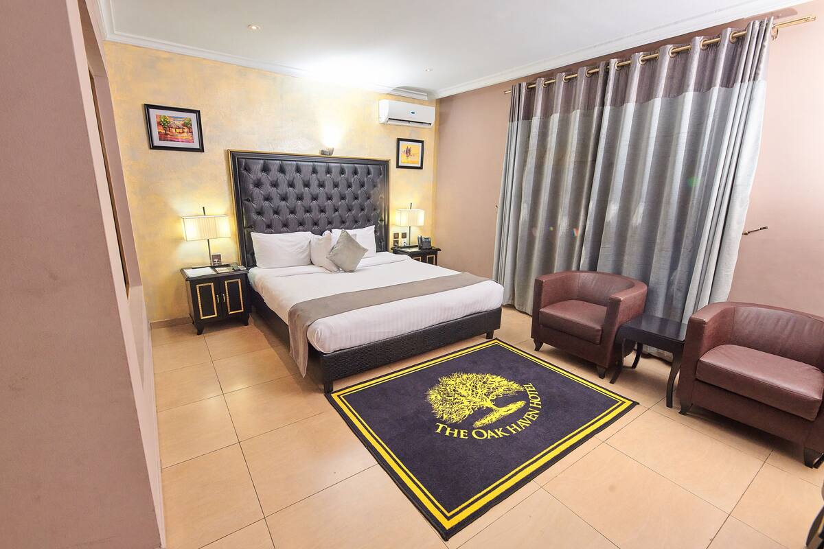 A hotel room with a large bed, two bedside tables, a nightstand, a sofa, and a chair. There is a rug with a logo on the floor.