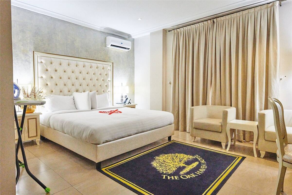 A hotel room with a large bed, a chair, a desk, an air conditioning unit, and a curtain.