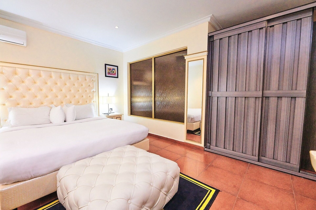 A hotel room with a large bed, a tufted headboard, a bedside table, a nightstand, a large mirror, and a wardrobe.