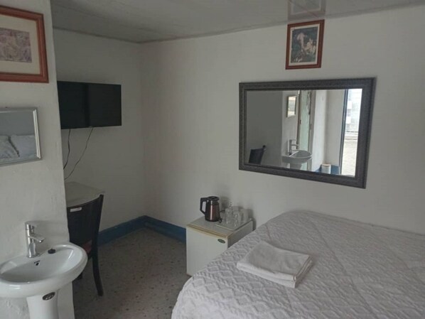 A hotel room with a bed, a bedside table with a kettle, a sink, and a mirror on the wall.