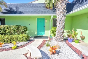 A green building with a turquoise door, surrounded by a garden with various plants and a palm tree.
