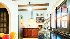 Une cuisine avec des meubles en bois, un plan de travail noir, un panier de fruits, une plante suspendue et une fenêtre avec des rideaux.