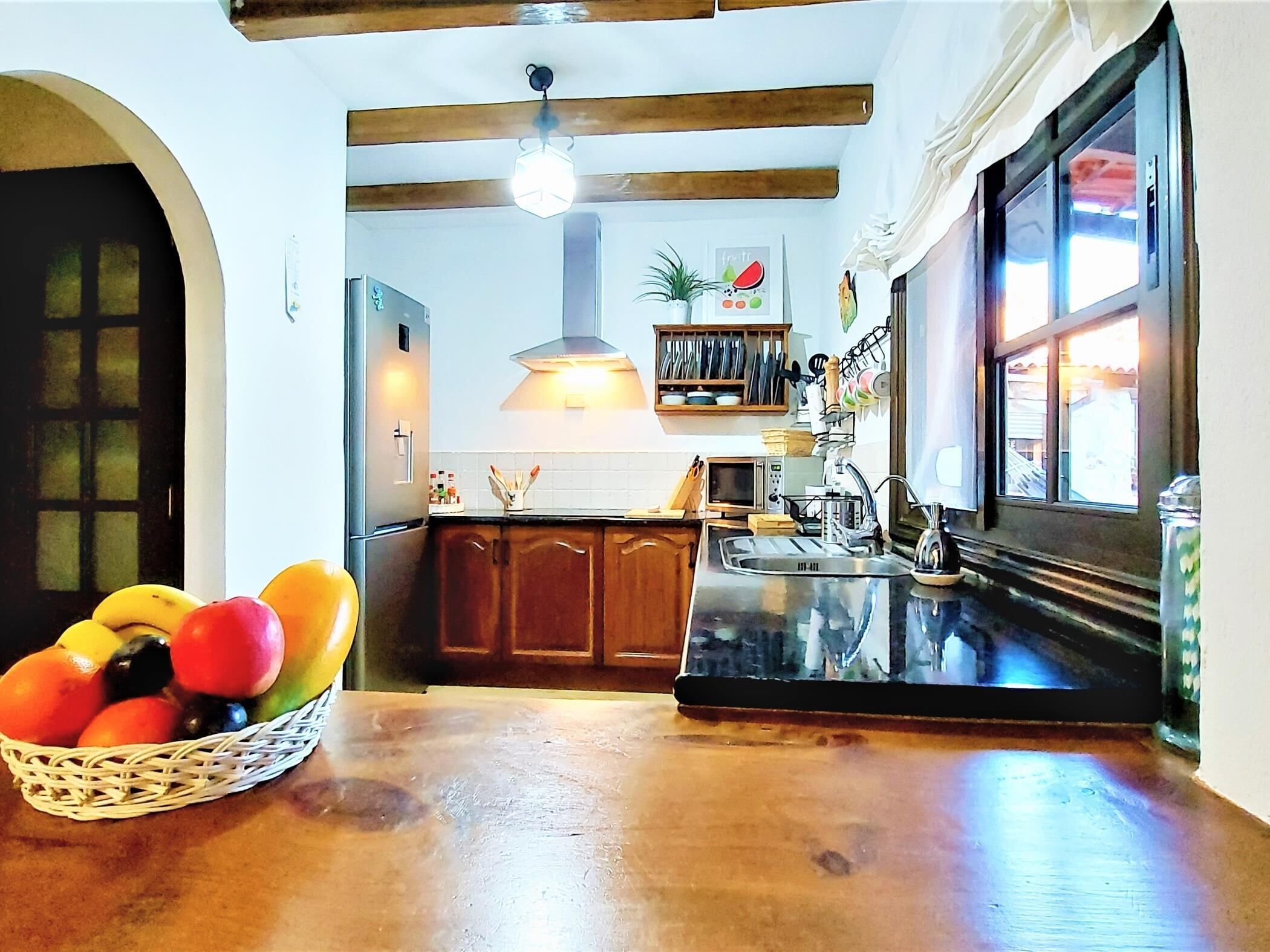 Een keuken met houten kastjes, een zwart aanrecht, een fruitmand en een raam met gordijnen.