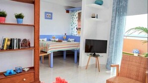 A room with a bed, a TV, a bookshelf, a dining table with a striped tablecloth, and a chair. - Beautiful holiday home with sea views +beach+WIFI (El Rosario)