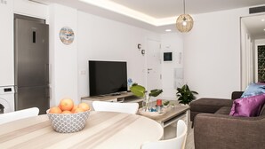 Una cocina moderna con refrigerador empotrado, una mesa de comedor con un bol de fruta, televisor de pantalla plana y una zona de estar con sofá y sillas.