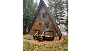 Una cabaña de madera con techo triangular, puerta principal y un pequeño porche con escalones.