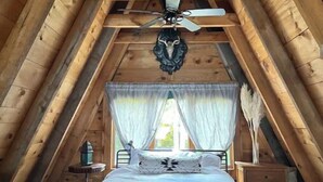 Un dormitorio estilo altillo acogedor con techo de madera, cama con ropa blanca, ventilador de techo decorativo y cortinas translúcidas.