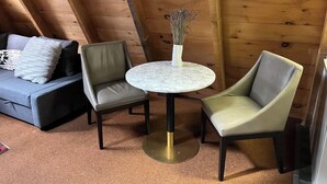Una habitación acogedora con techo de madera inclinado, un sofá gris con cojines estampados, dos sillones grises y una pequeña mesa de mármol con un jarrón de plantas secas.