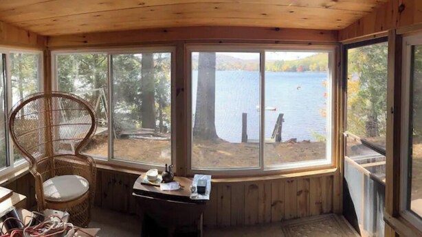 Una cabaña de madera con amplios ventanales que dan a un lago, una silla de mimbre y una mesa con objetos encima.