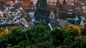 Uma catedral histórica com uma torre alta, rodeada por um horizonte urbano ao pôr do sol.